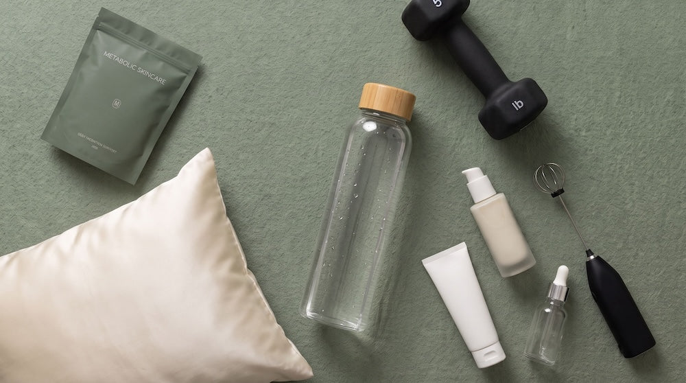 Flat late image of objects that relate to the article - Metabolic Skincare package, silk pillow case, water bottle, serums, weights, and a whisk